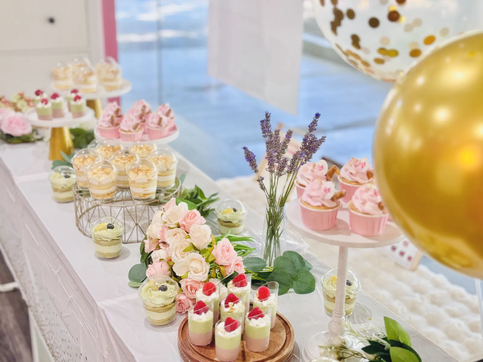 A styled dessert table with pink cupcakes, dessert cups, roses and gold balloons
